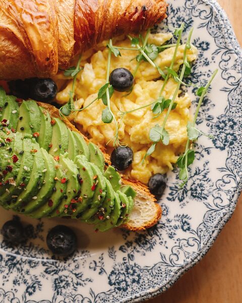 Croissant abierto con huevo revuelto, aguacate en abanico y arándanos, en plato de loza azul