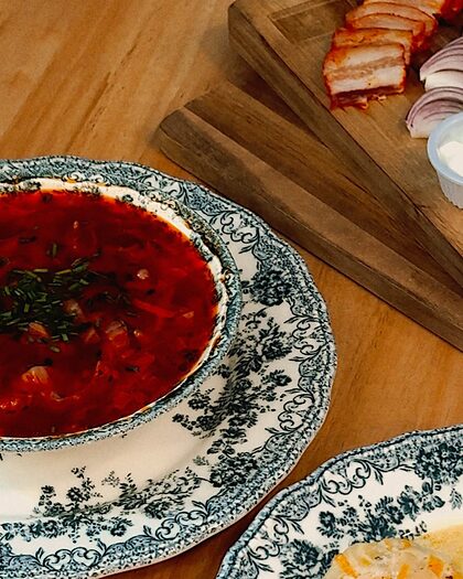 Plato hondo de borsch con cebollino en loza azul, junto a una tabla con salo, cebolla morada y pan negro