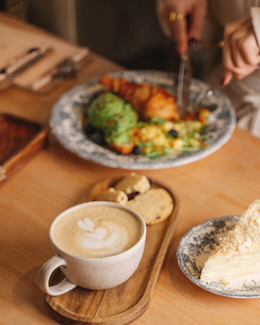 Mesa de madera con desayuno, capuchino y manos compartiendo el plato