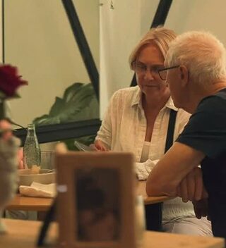 Una pareja mayor conversa en una mesa junto a la ventana