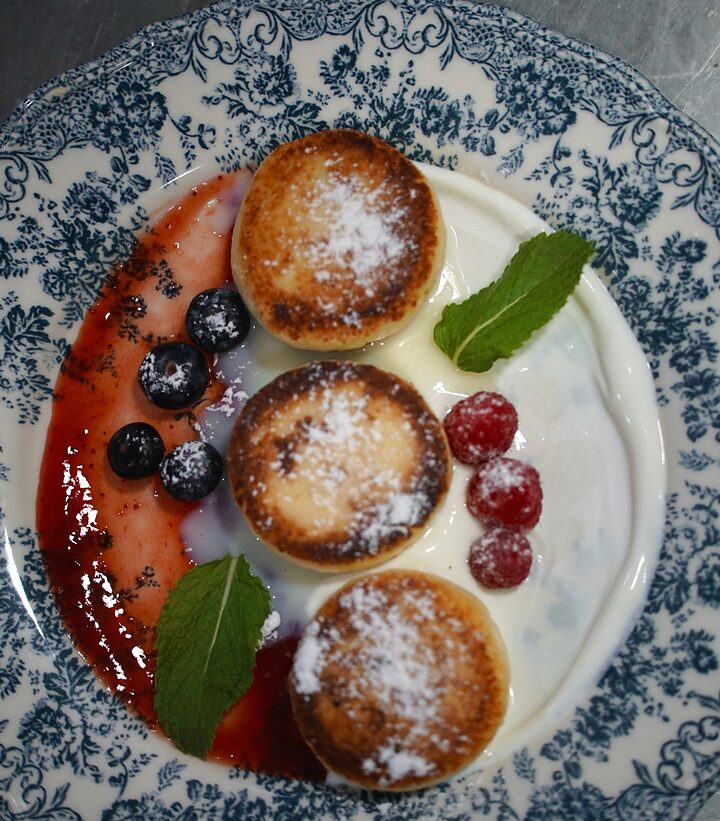Tres syrniki con azúcar glas sobre crema agria y frutos rojos, en un plato de loza azul
