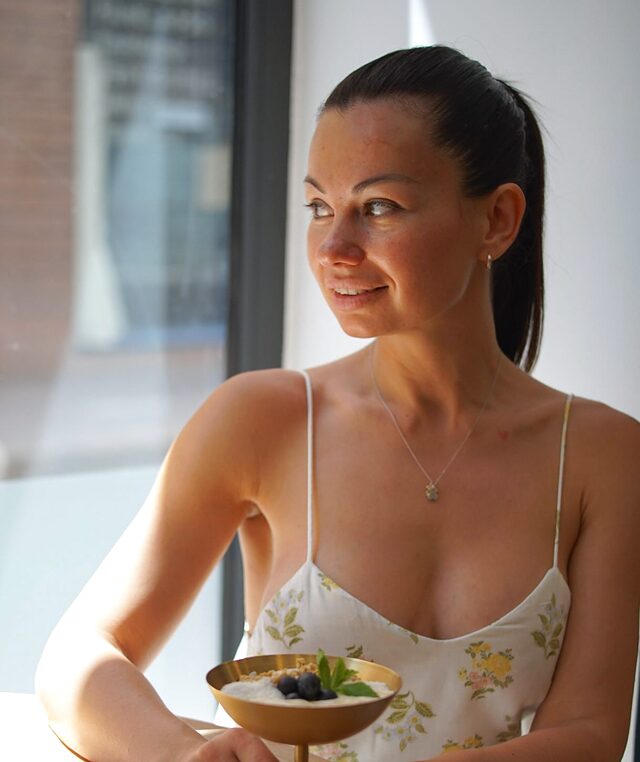 Una chica junto a la ventana con un cuenco de postre en la mano, sonriendo